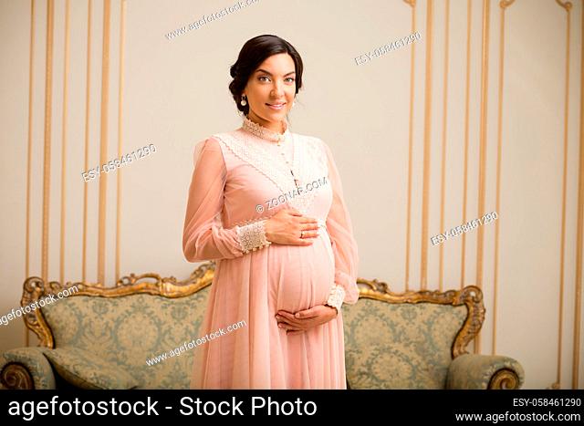 Picture of happy brunette pregnant lady in pink dress posing in vintage interior in expensive apartment. Royalty concept