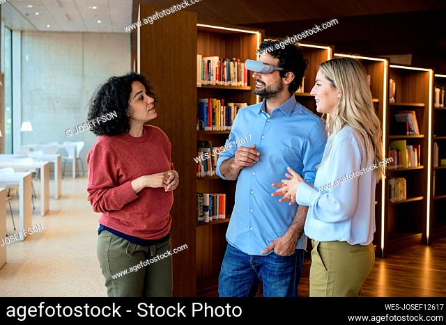 Smiling man wearing futuristic glasses talking with colleagues in library