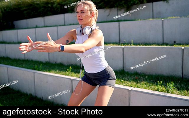 Blond sportswoman with headphones stretching out arms and squatting near grassy steps during fitness training in park on summer day