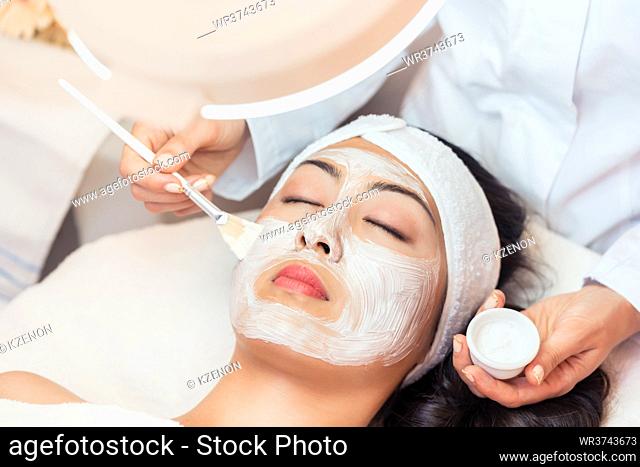 Close-up of the face of a young woman relaxing under the gentle touch of the specialist, applying on her cheeks white facial mask with rejuvenating effects