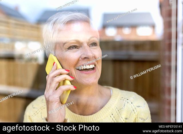 Happy mature woman talking on smart phone while looking through glass window