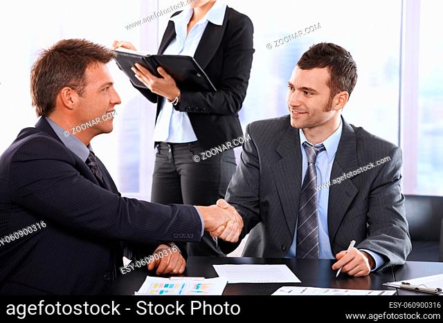 Businessmen shaking hands at meeting, sitting at table, assistant in background holding organiser