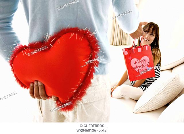 Mid section view of a man hiding a heart shaped cushion and giving a gift to a young woman