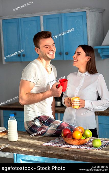 Laughing couple in at the kitchen in the morning. Man and woman smiling, talking and holding cups with coffe in their hands