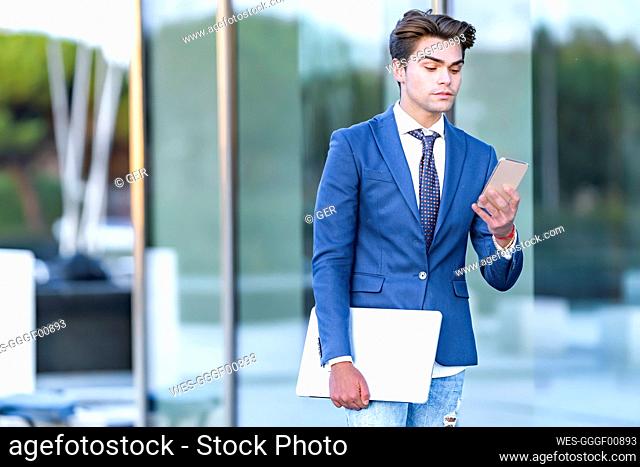 Young businessman with laptop using mobile phone against office building