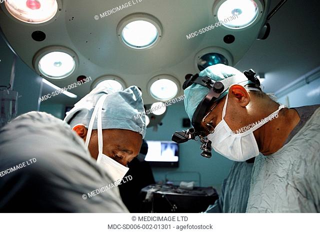 A heart surgeon and his assistant operate on a patient. The lead surgeon wears magnifying lenses over his glasses. The magnifying lenses will give the surgeon a...