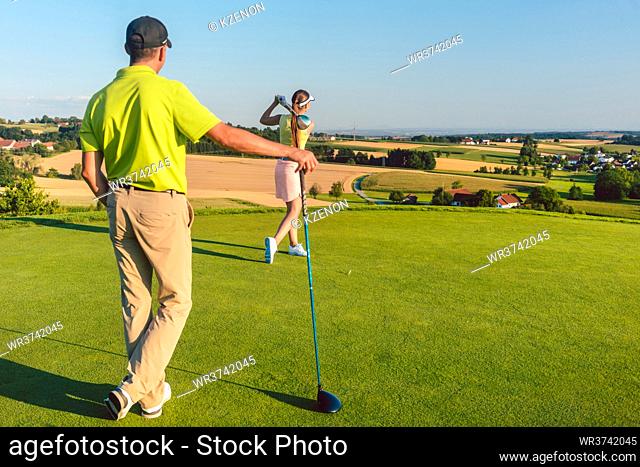 Full length rear view of a man wearing modern golf outfits, while watching his partner striking the ball during match on the green grass of a professional golf...