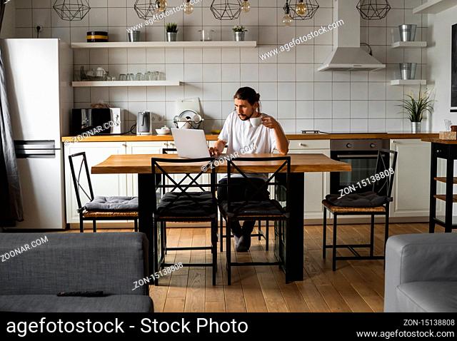 Freelancer working from home on the kitchen and using laptop. Handsome smiling man holding cup of coffee and looking at laptop