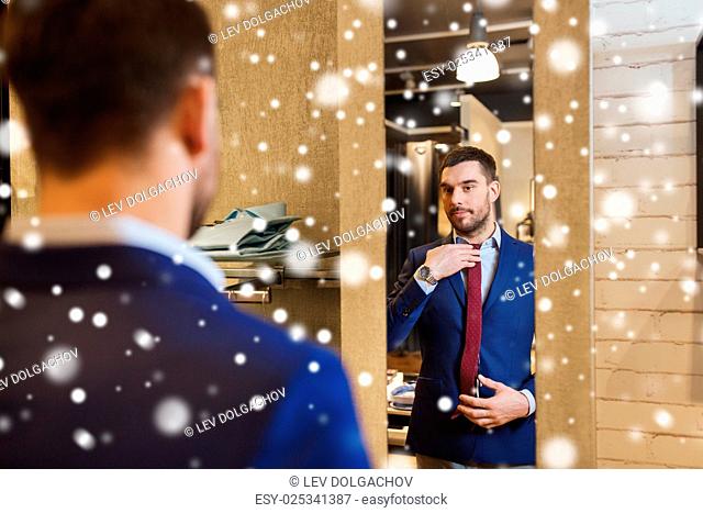 sale, shopping, fashion, style and people concept - elegant young man choosing and trying jacket on and looking to mirror in mall or clothing store over snow