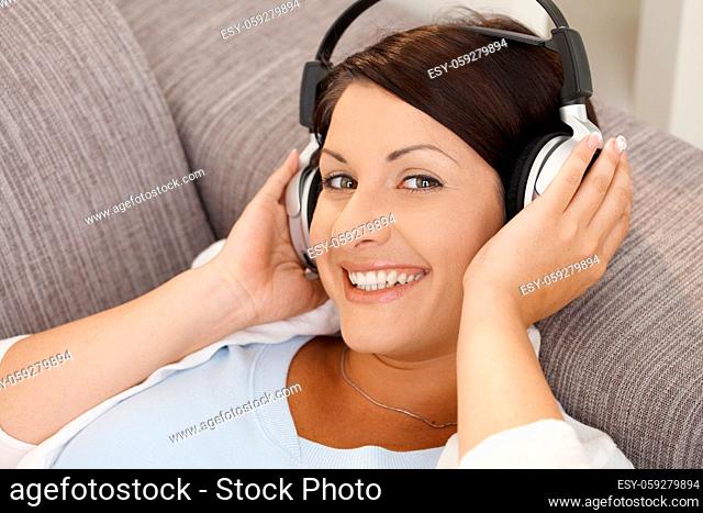 Happy young woman listening music on headphones, lying on sofa at home, smiling