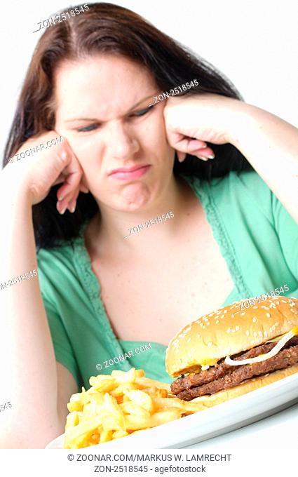 erwachsene übergewichtige Frau mit schwarzen Haaren sitzt am Tisch und kämpft mit einem Hamburger und Pommes Frites, vor weißem Hintergrund
