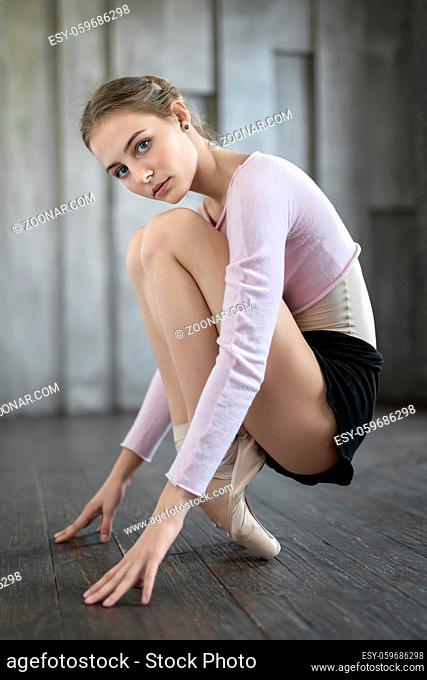 Sensual ballerina sits on the toes and holds hands on the floor on the blurry wall background. She wears black shorts, light leotard with lilac blouse and...