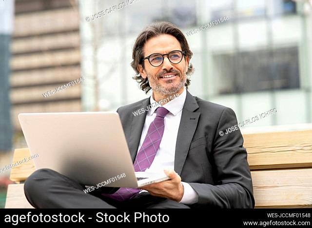 Smiling male entrepreneur with laptop looking away while sitting on bench
