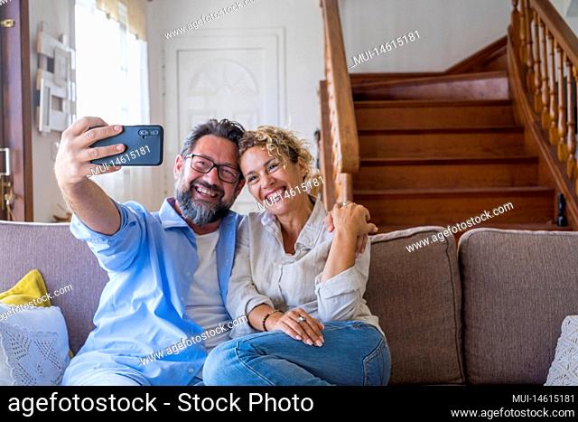 Happy caucasian couple taking selfie sitting on sofa using mobile phone camera at home. Joyful wife and husband taking self portrait on smartphone