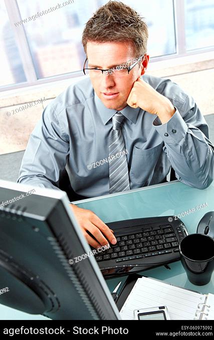 Handsome mid-adult businessman deep in thought concentrating on computer work looking at screen sitting at office desk