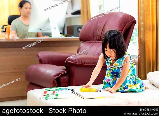 Asian girl child reading interactive book in living room at home as home schooling while her mom working at home because city lockdown because of covid-19...