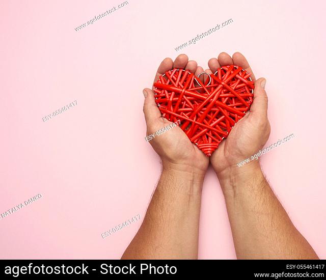 two male hands holding a red wicker heart on a pink background, love concept, copy space
