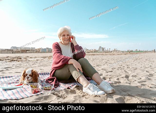 Pleased elderly lady sitting on the sand beside her cute pet during the phone talk
