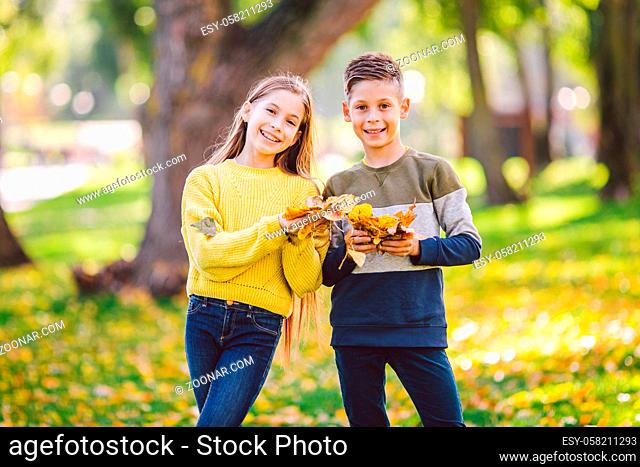 Happy twins teenagers boy and girl posing hugging each other in autumn park holding fallen yellow leaves in hand in sunny weather. Autumn season theme