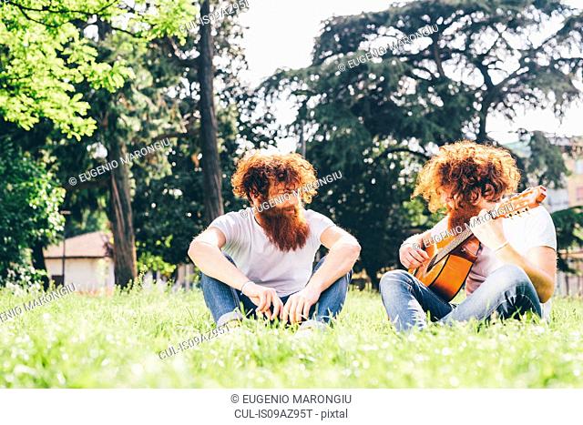 Young male hipster twins with red beards sitting in park playing guitar