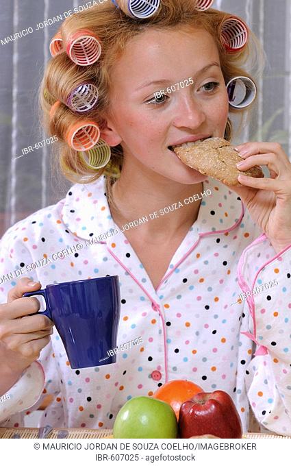 Redheaded woman wearing pajamas with curlers in her hair eating and drinking from blue cup