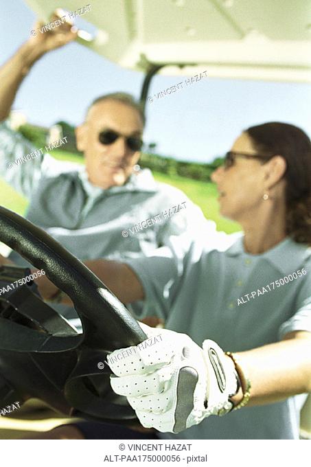 Mature man and woman in golf cart, close-up, focus on hand and steering wheel in foreground