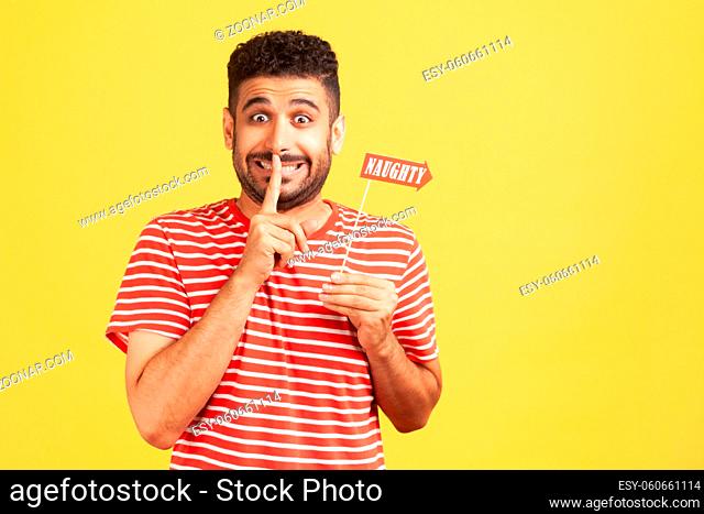 Funny devious bearded man in striped t-shirt holding finger near lips showing shh gesture holding in hand stick with naughty inscription, surprise