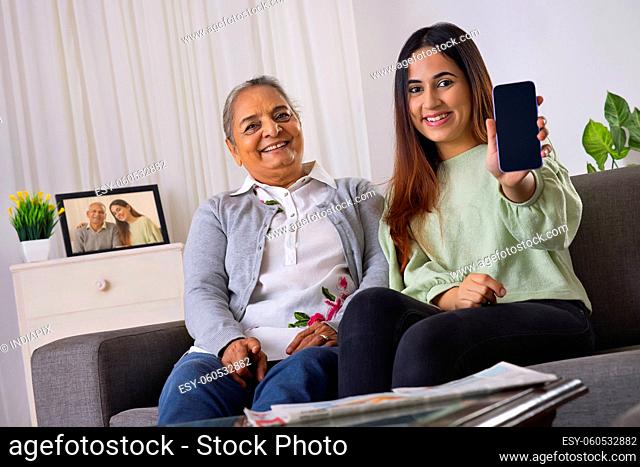 Young lady with her grandmother sitting on sofa and showing smartphone