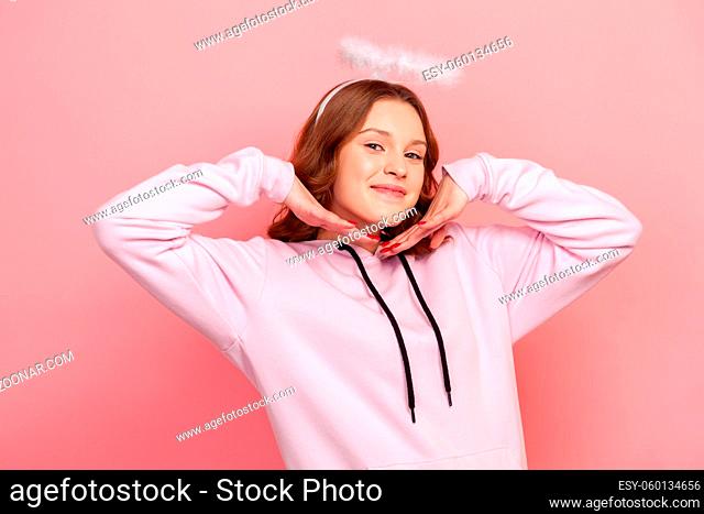Portrait of cute smiling curly haired teenage girl in hoodie with halo on her head posing on camera with happy face, I am angel gesture
