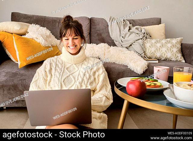 Smiling woman in sweater using laptop while sitting in living room at home