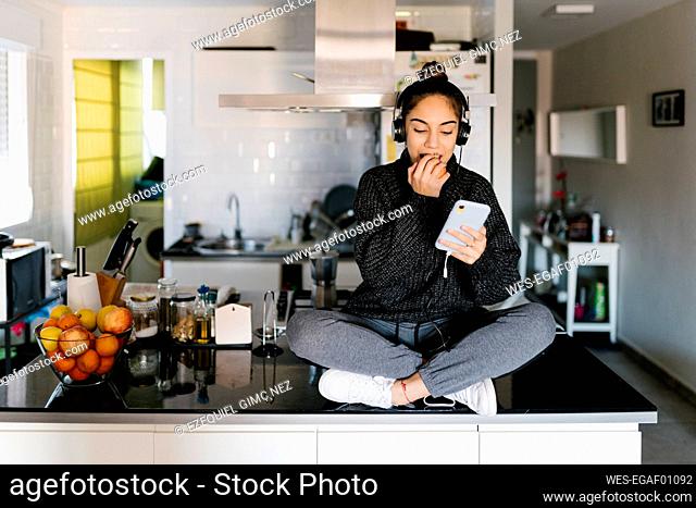 Teenage girl wearing headphones eating fruit while using mobile phone sitting at home