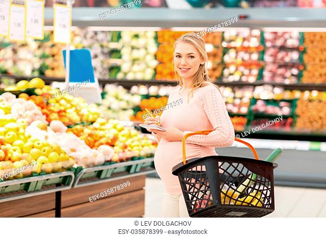 sale, shopping, food, pregnancy and people concept - happy pregnant woman with basket and notebook at grocery store or supermarket