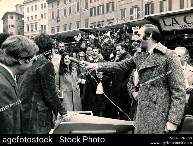 Italian TV host Pippo Baudo interviewing some people in the street market. Italy, 1970s