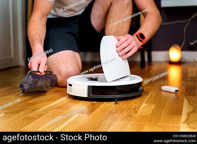Caucasian young man doing white robot vacuum cleaner maintenance. Autonomous wireless modern vacuum cleaner. Male repairing robot cleaner home on floor