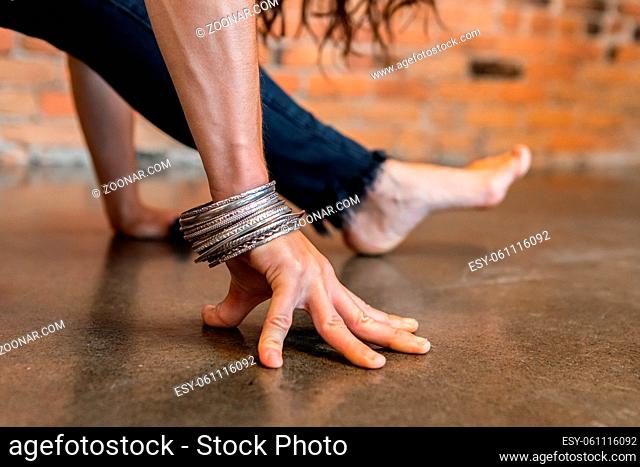 A close up view on the hand of a woman wearing silver bracelets during a Vinyasa yoga workout, in a leg stretch pose with hands resting on floor