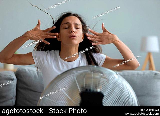 Woman seated on couch in living room suffers from swelter waving her hands trying to cool off by the fan, oppressive heat too hot weather and sultriness in...