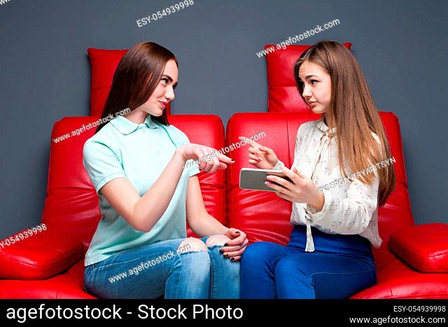 Two girlfriends sitting on red leather couch and looking at pictures on phone