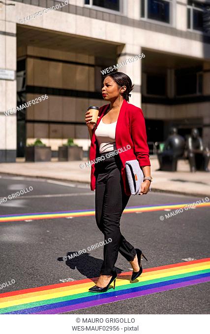 Businesswoman with coffee to go and laptop bag crossing the street on LGBT stripes, London, UK