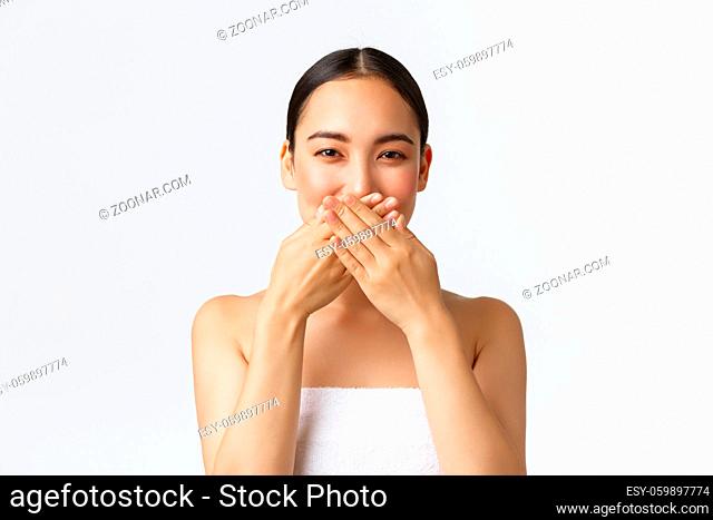 Beauty, cosmetology and spa salon concept. Beautiful asian woman in towel hiding lips, cover mouth with hands and giggle silly, smiling with eyes
