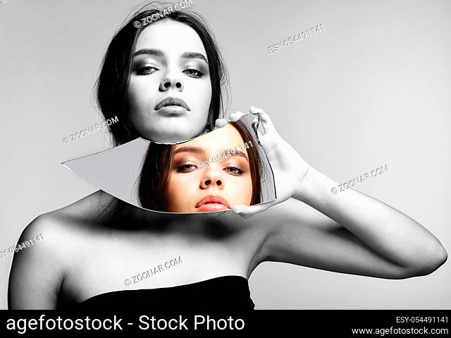 Monochrome portrait of girl with a shard of the mirror. Female with mirror shard in hand posing on gray background. Color face reflection in mirror splinter