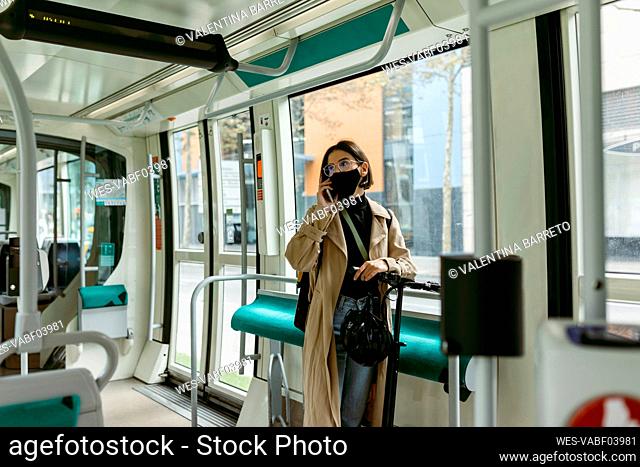 Woman with electric push scooter and face mask talking on mobile phone while standing in tram