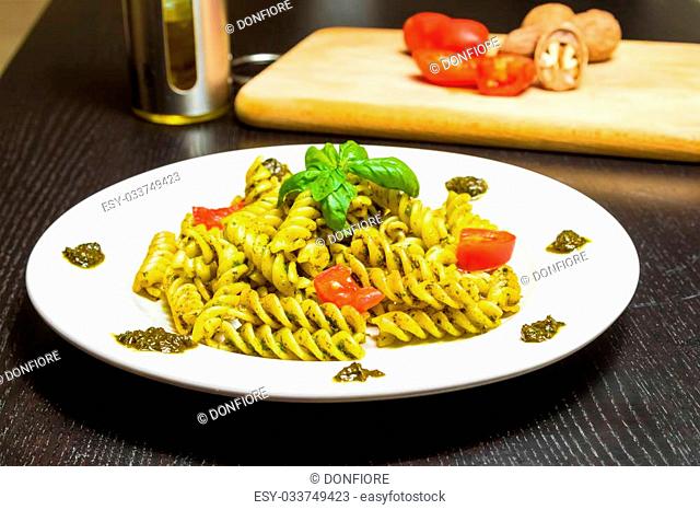 dish of pasta with pesto genovese sauce and vegetables on black wood table, with ingredients, walnuts, tomato, basil and olive oil in background