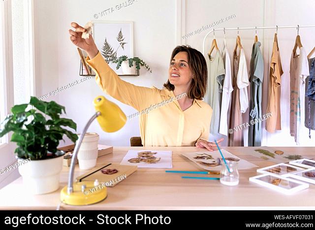 Smiling beautiful analyzing dry leaves while sitting at table against clothes rack