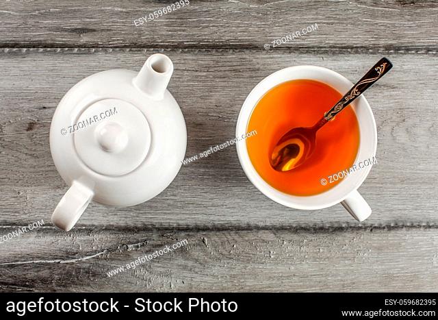 Table top view on small white teapot and cup full of hot amber tea with silver spoon in it, placed on gray wood table