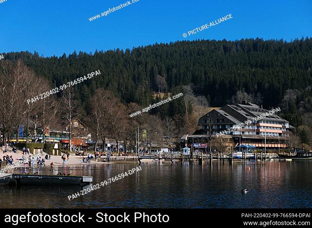 PRODUCTION - 27 March 2022, Baden-Wuerttemberg, Titisee-Neustadt: A hotel stands on the shore of Lake Titi while tourists can be seen next to it on the shore