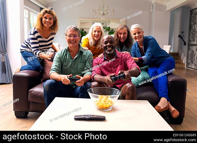 Multiracial senior male friends playing video game while women cheering them in background at home
