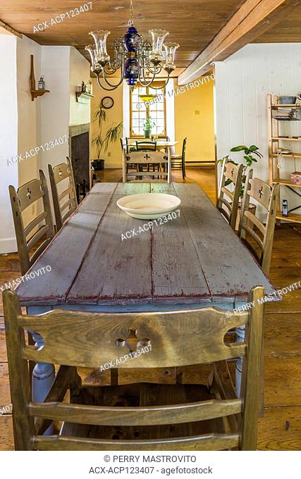 Wide plank wooden dining table with empty white ceramic bowl and high back chairs in dining room inside an old circa 1760 Canadiana fieldstone cottage style...