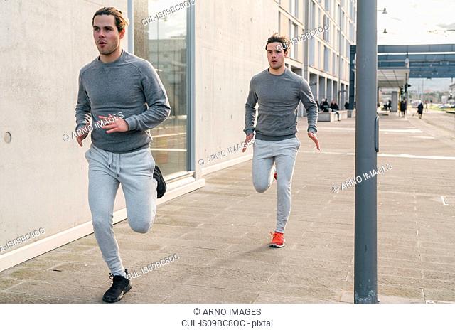 Young adult male twin runners, running along city sidewalk