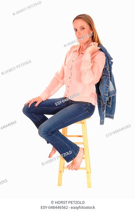 A young slim woman sitting bare feet on a chair in jeans with her jeans.jacket over her shoulder, isolated for white background