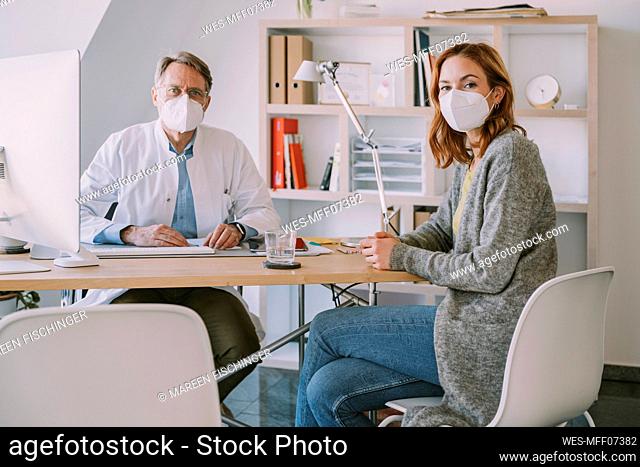 Doctor and female patient wearing protective face mask sitting by desk at doctor's office